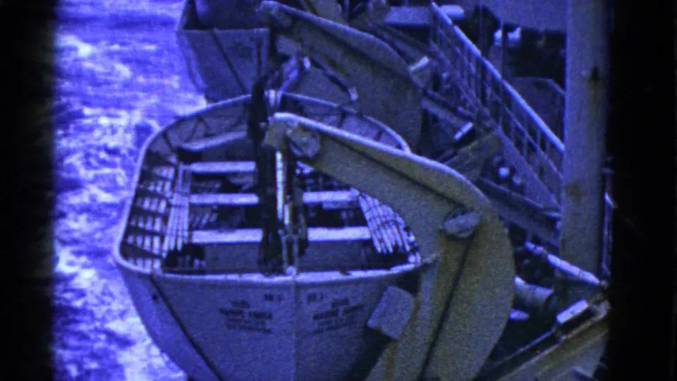 Shipboard view of a lifeboat held in davits, with blue-tinted water and deck fittings visible in the frame.
