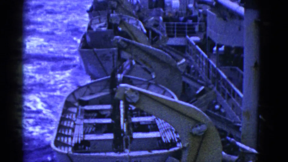 Blue-tinted 8mm frame showing multiple lifeboats arranged along a ship deck with davits and railings.
