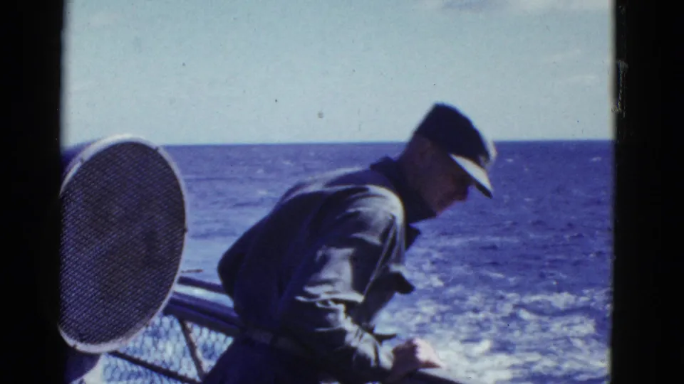 A uniformed man aboard a vessel near San Francisco leans beside the rail, with ocean, wake, and a circular mesh fixture behind him.