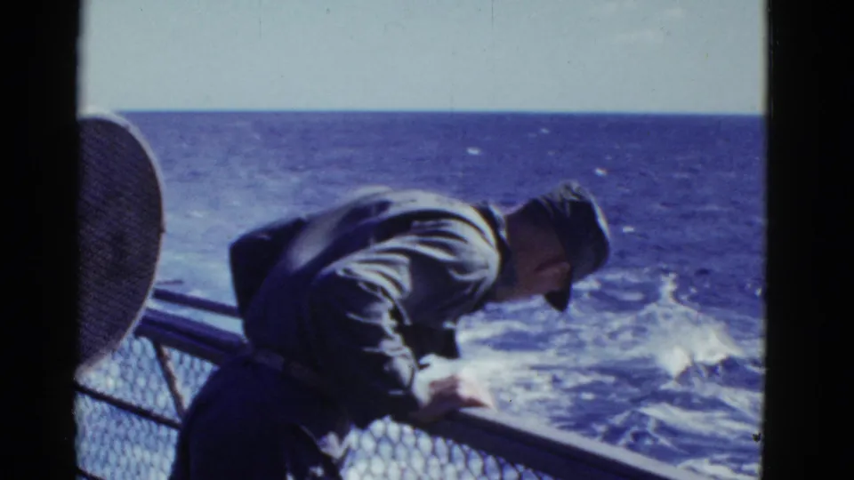 A uniformed man on a vessel near San Francisco bends over the rail and looks down toward the moving water below.