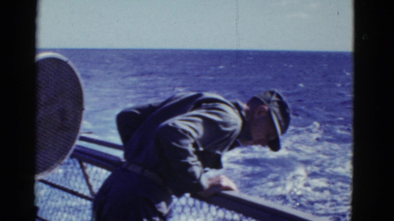 Uniformed man leans over a ship railing with open ocean and deck equipment behind him.