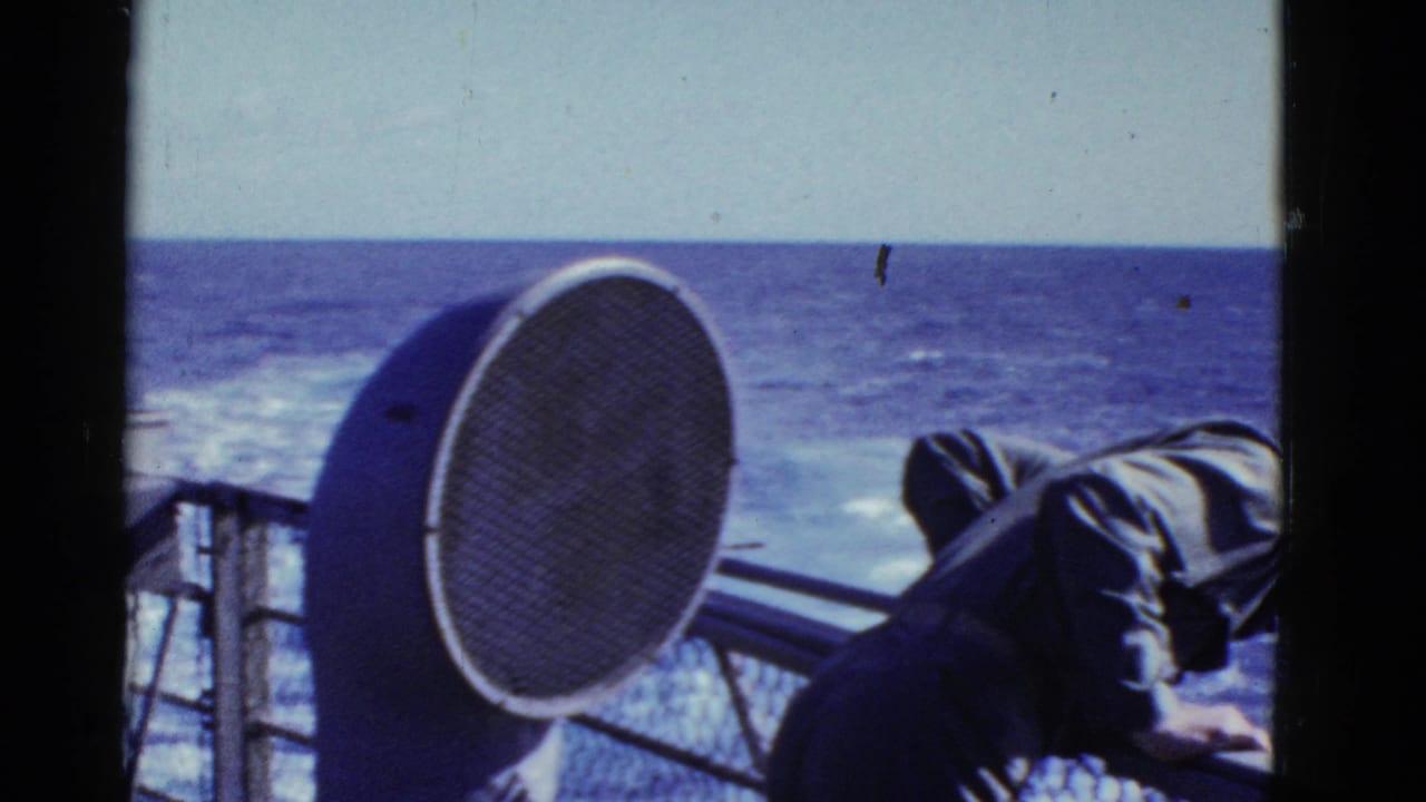 A dark-uniformed man is partly visible at the rail, with blue ocean, wake and a circular deck speaker in view.