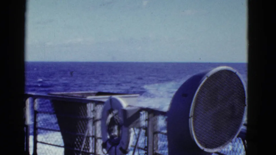 A ship deck view shows ocean wake, railings, a life preserver and speaker, with the man cropped near the right edge.