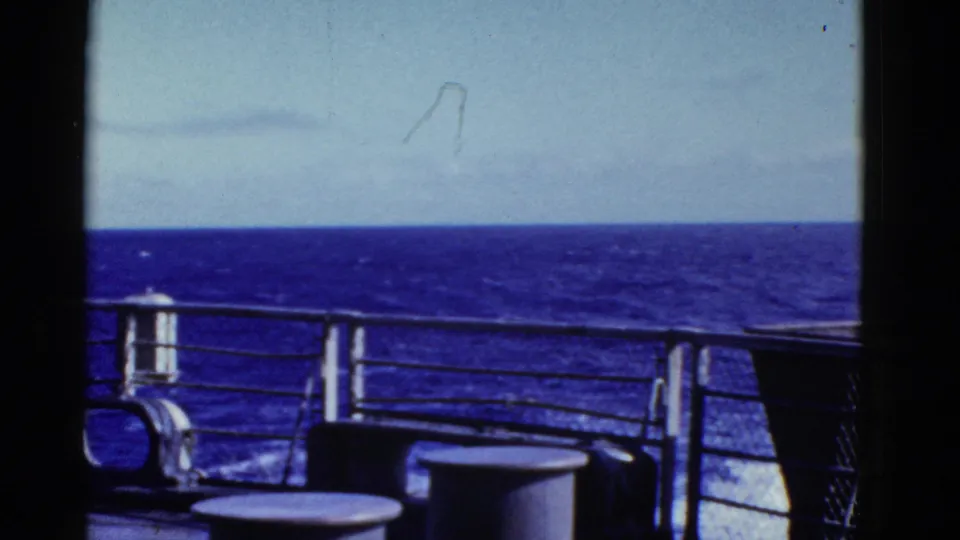 The pan continues across ship railings and deck fittings toward a calm horizon, with 8mm grain and visible film edges.