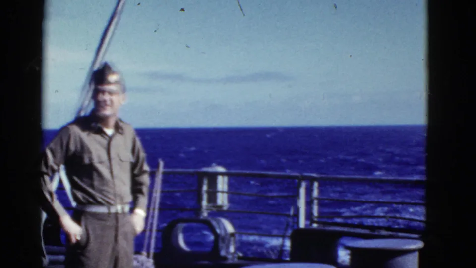 Uniformed adult man smiles toward the camera on a ship deck, with railings and open ocean behind him in the 1952 8mm footage.