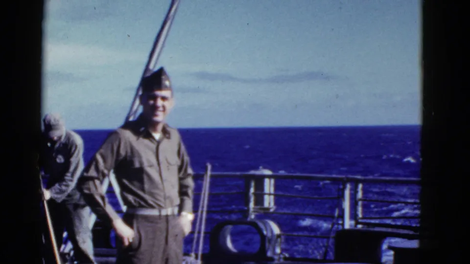 Early-1950s 8mm frame of a uniformed man smiling on a ship deck at sea, with another man working near the rail.