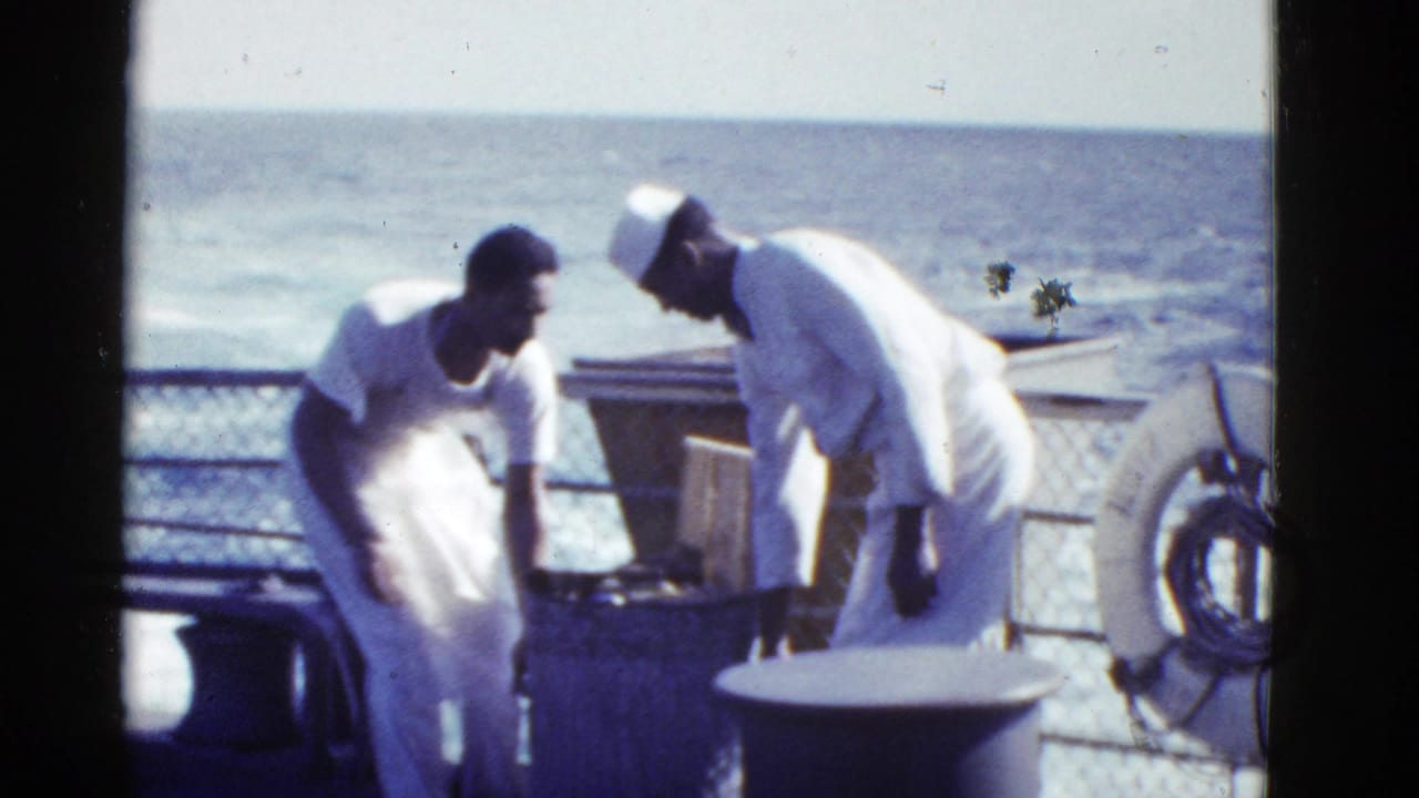 Two sailors in white uniforms stand near deck equipment and a life ring at sea.
