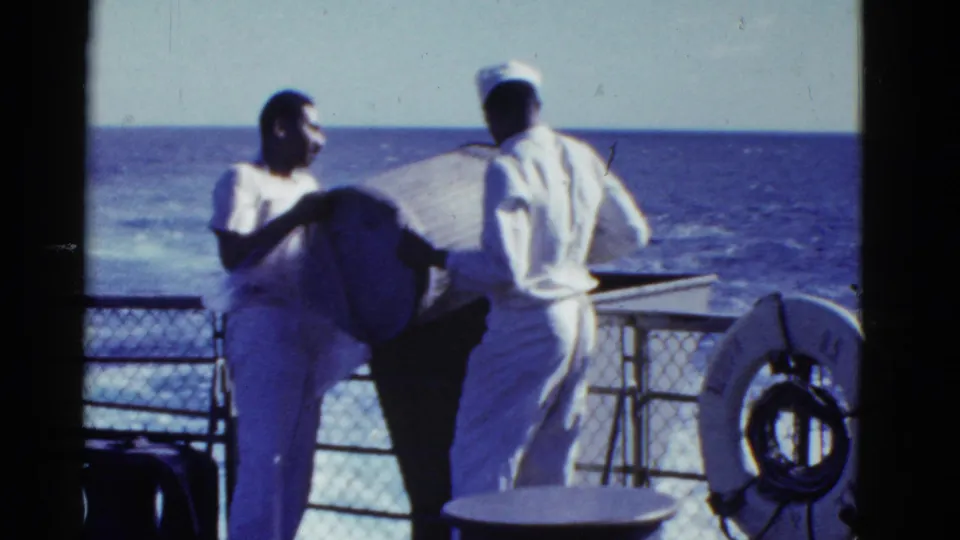 1950s home-movie frame of two sailors on deck near a railing and life ring, with open sea in the background.