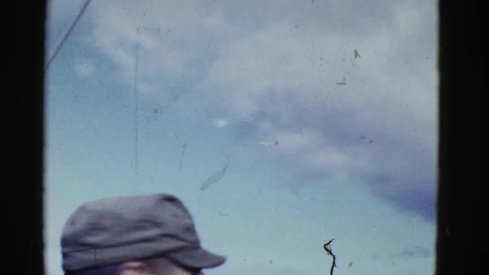 1950s 8mm frame looking up at a cloudy San Francisco sky, with the crown and brim of a cap at the bottom.