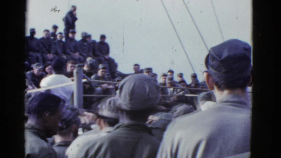 A handheld 8mm view shows servicemen in caps and jackets watching a shipboard boxing match in the early 1950s setting.