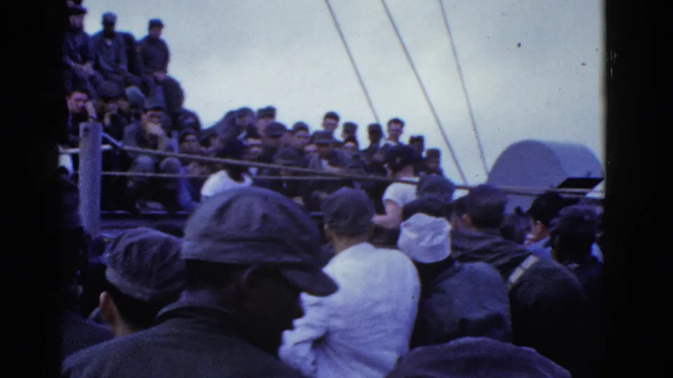 Color 8mm frame of a shipboard crowd, with adult men in caps and uniform-like clothing watching activity on deck.