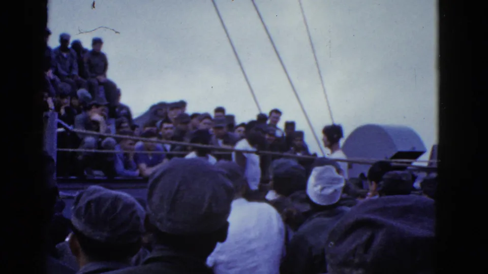 Handheld view from within a shipboard crowd, with foreground heads and caps facing a deck activity beyond the railing.