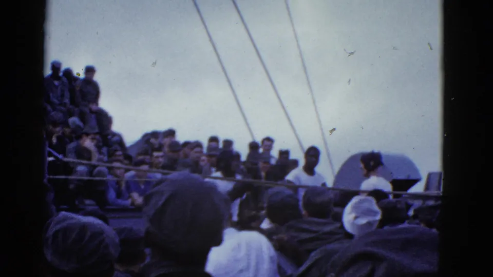 Wider 8mm frame of a vessel deck crowd, rigging, and a rounded deck structure as spectators face a shipboard event.