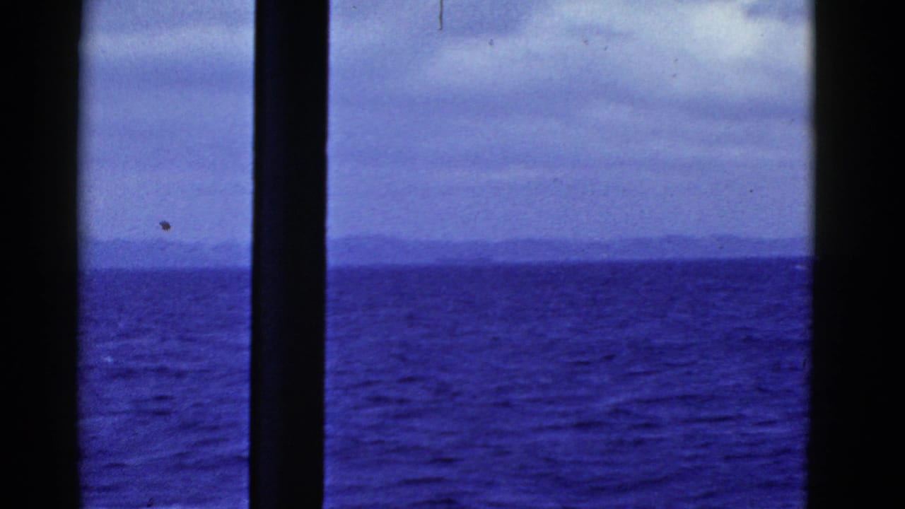 1952 San Francisco home-movie frame showing blue ocean, distant land, and cloudy sky through dark ship-window mullions.