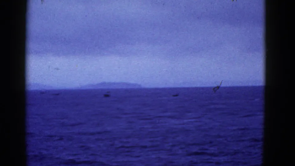 Blue-purple 8mm frame of San Francisco Bay-area water with small distant boats and low land visible through a dark cabin-window opening.