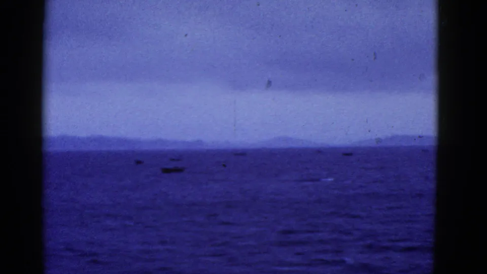 Blue-tinted 8mm frame of small boats on open water near San Francisco, with distant landforms under an overcast sky.