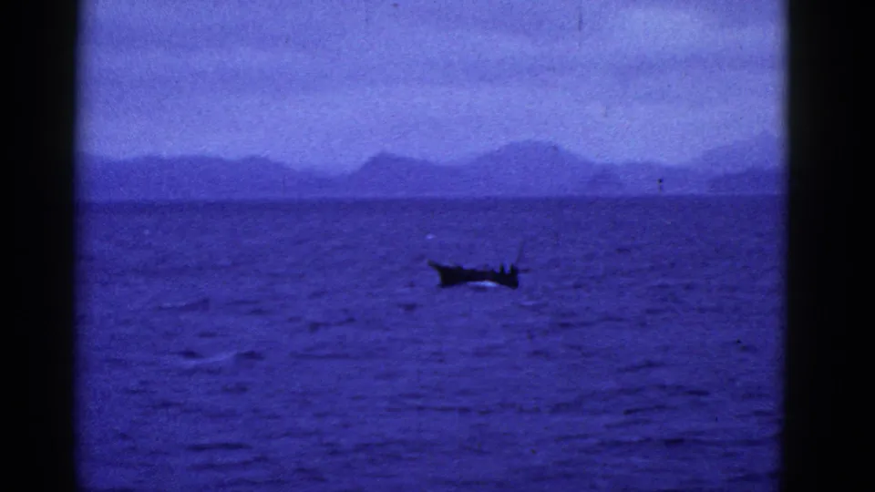 1952 8mm footage frame with a distant small boat on rippled blue water, backed by a pale sky and mountainous coastline.