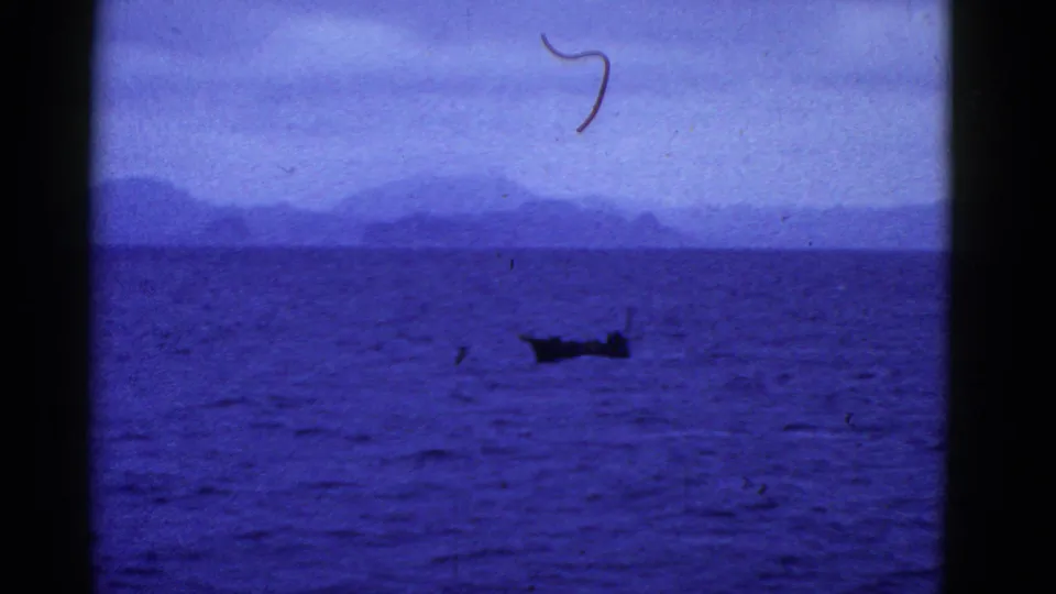 The 1950s 8mm frame holds on a small boat across deep blue water, with distant landforms softened by haze.