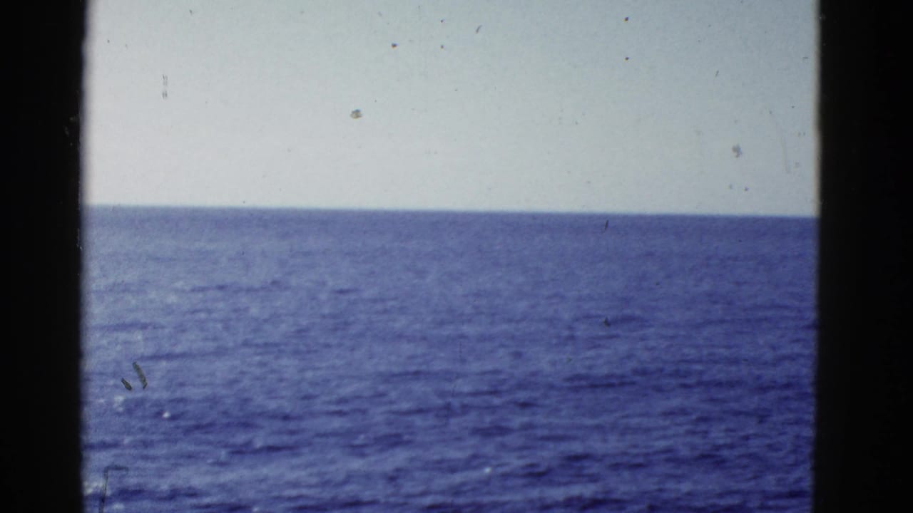 1950s home-movie view of calm blue ocean meeting a pale sky, with dark vertical edges suggesting a vessel-window perspective.