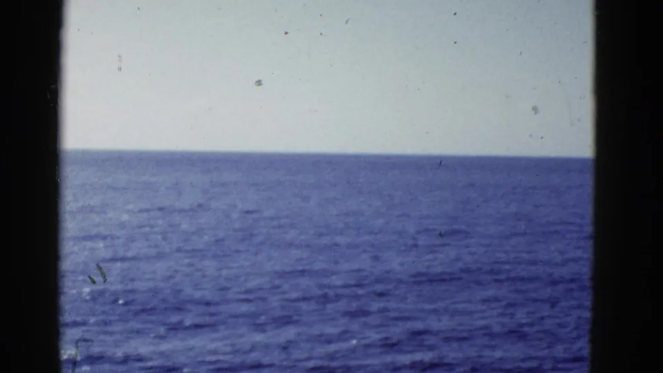 1950s home-movie view of calm blue ocean meeting a pale sky, with dark vertical edges suggesting a vessel-window perspective.