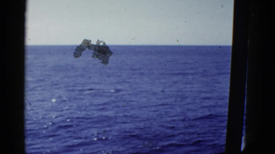 Blue ocean and pale sky in a 1950s home-movie frame, with a dark vertical edge and a larger film mark over the water.