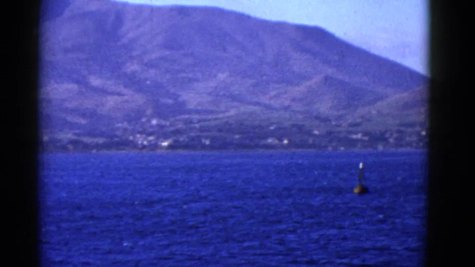 1952 8mm view from a boat on San Francisco Bay, with blue water, a buoy, and a rugged island or shoreline in the distance.