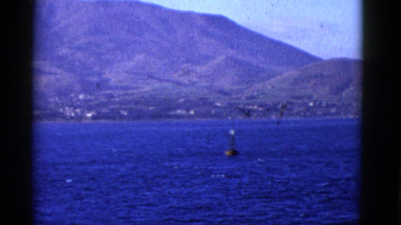 Mountainous shore across blue water with a buoy near the center of the frame.
