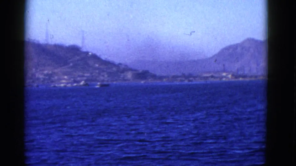 1952 bay travel footage showing water, a low shoreline, hazy hills, and slender towers in the distance.