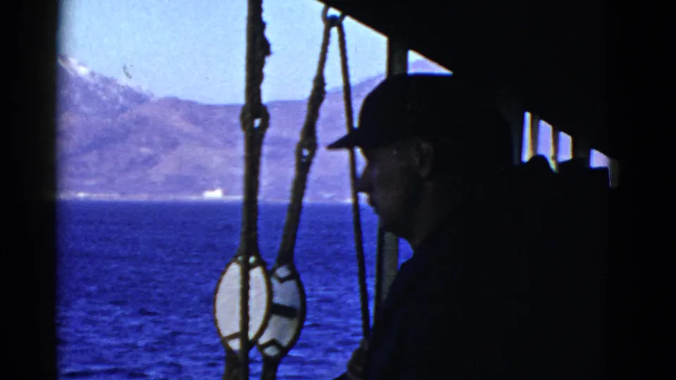A silhouetted adult man in a cap stands on a ship deck, with ropes, blue water, and a hazy shoreline behind him.