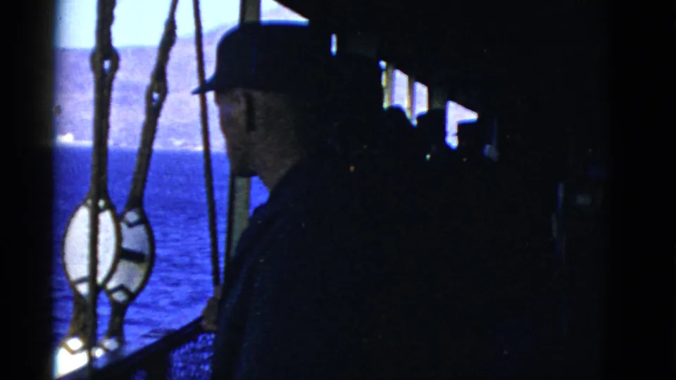 An adult man in a cap remains in shadow on a ship deck, looking out toward blue water and a distant San Francisco shoreline.