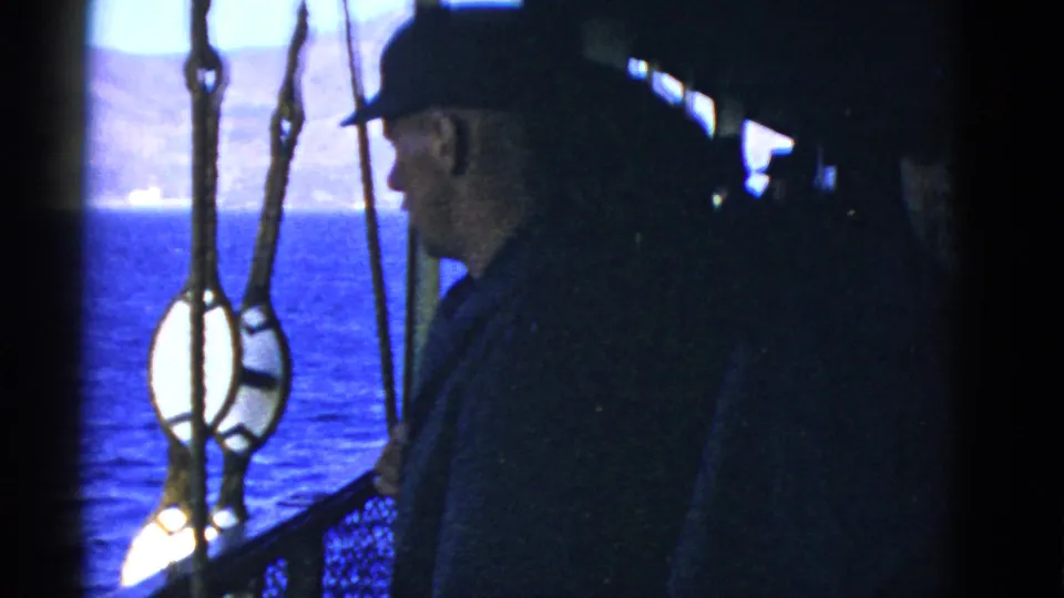 1950s 8mm view from a ship deck showing a man near rigging with blue ocean and a mountainous shoreline beyond.