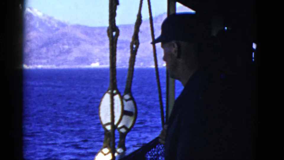 A man in a dark cap stands at the edge of a shaded ship deck, with blue water and distant mountains visible beyond.