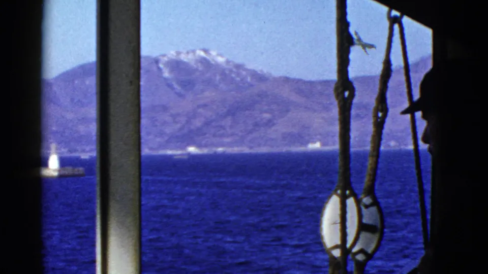 From a boat, the frame shows a silhouetted man, rigging, blue water, mountains, and a small lighthouse or beacon near the lower left.