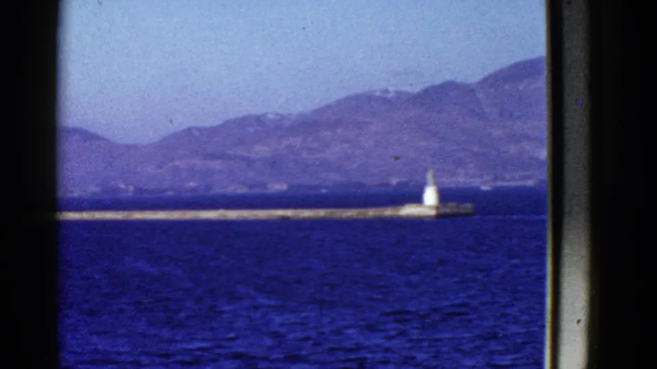 Early 1950s San Francisco Bay shoreline view with deep blue water, a low pier, white beacon, and hazy mountain ridge framed by vessel structure.
