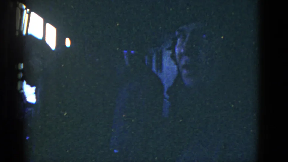 Inside a dark vessel interior, a silhouetted man appears to hold a camera near bright windows, with other forms in shadow.