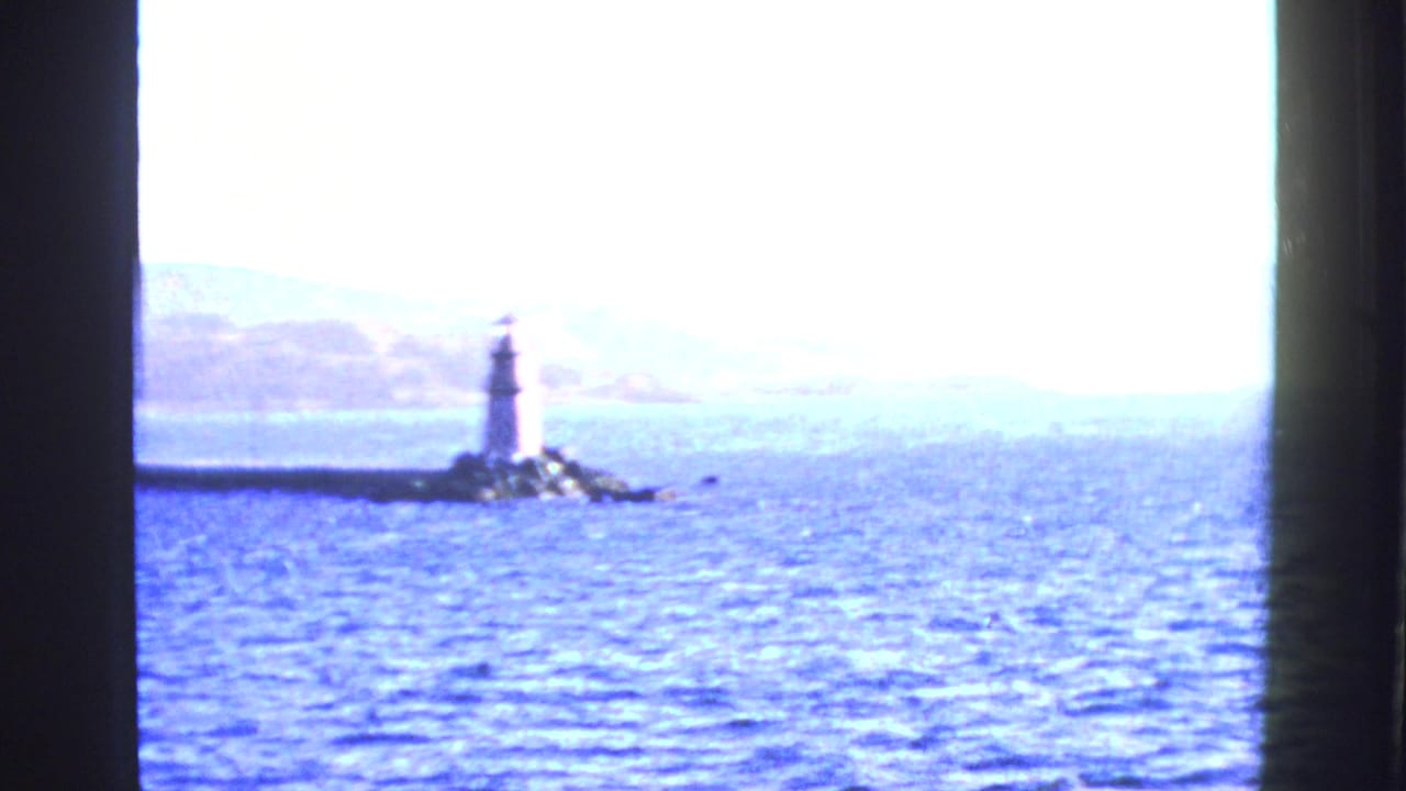 1950s San Francisco Bay home-movie frame showing a lighthouse or beacon on rocks, bright water, distant hills, and dark window-like bars.