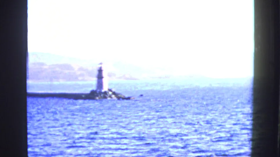 1950s San Francisco Bay home-movie frame showing a lighthouse or beacon on rocks, bright water, distant hills, and dark window-like bars.