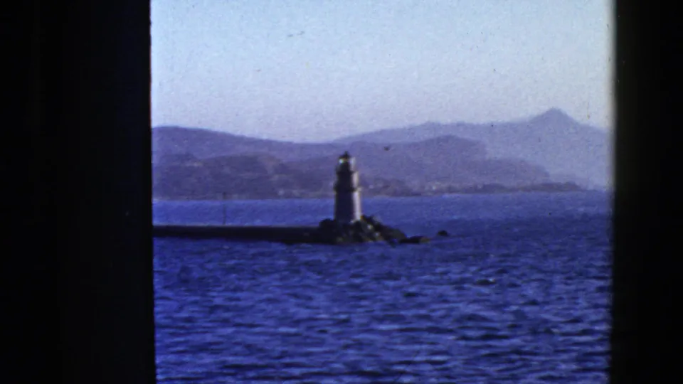 1950s San Francisco Bay view of a white lighthouse or beacon on rocks, seen beyond dark vertical bars with low hills across the water.