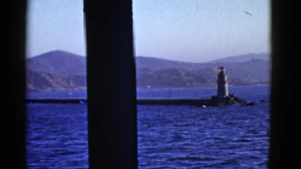 1950s home-movie frame from San Francisco Bay showing a lighthouse on a rocky outcrop, rippled water, distant hills, and dark vertical framing.