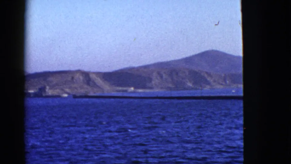 1950s 8mm coastal view with low hills, blue bay water, and dark framing from a window or railing.