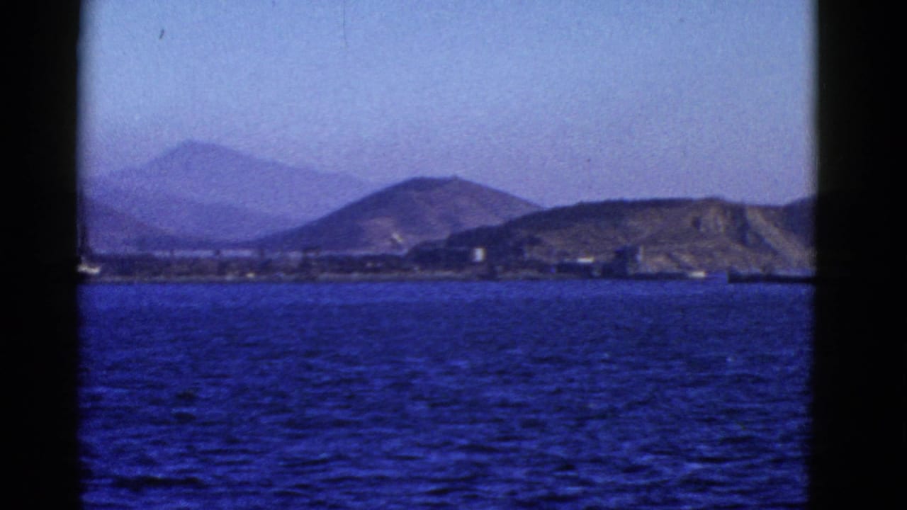 1952 San Francisco harbor view from the water, with blue bay surface, distant hills, shoreline buildings, and dark framing from the filming position.