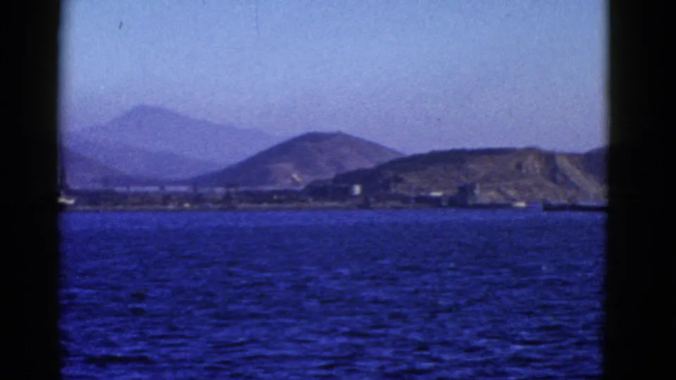 1952 San Francisco harbor view from the water, with blue bay surface, distant hills, shoreline buildings, and dark framing from the filming position.
