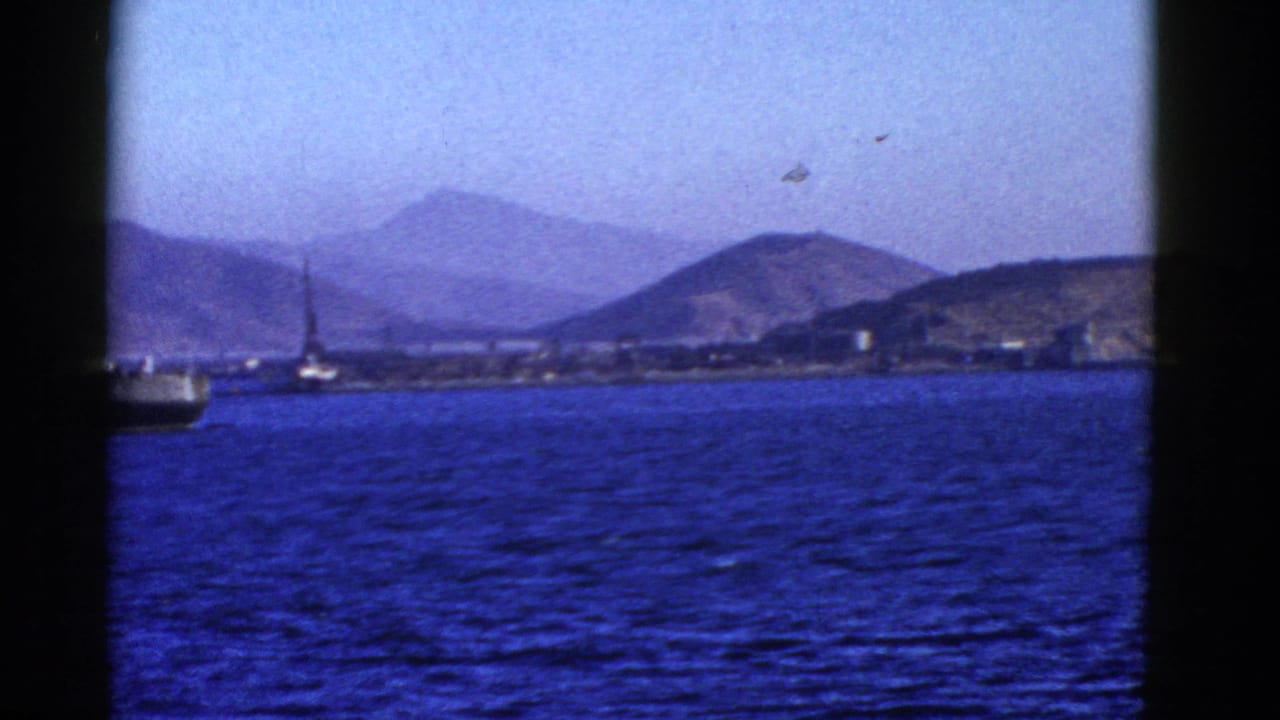 Harbor water with a distant shoreline, hills, and small vessels near a dock or crane.
