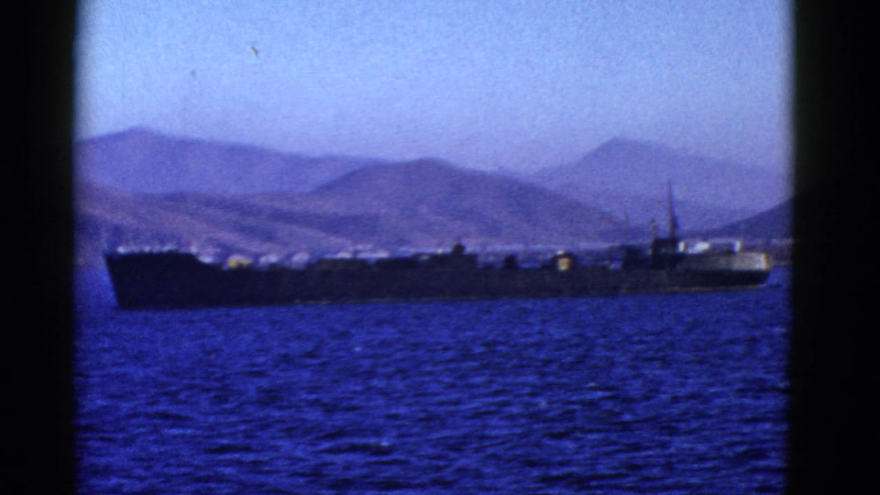 Cargo ship silhouetted on the water with mountains and shoreline in the distance.