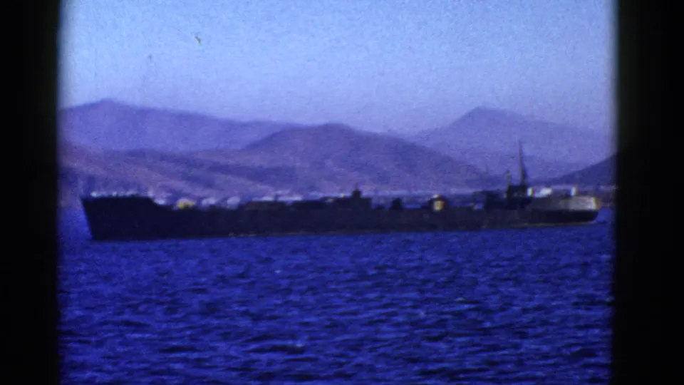 The vessel appears in a darker silhouette against deep blue water, with hazy coastal mountains beyond it in the 1950s home-movie frame.