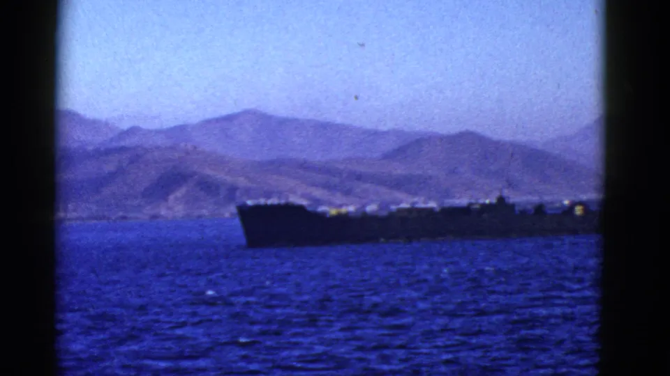 A dark ship is partially framed along the lower edge of the shot, with blue water and distant mountains visible behind it.