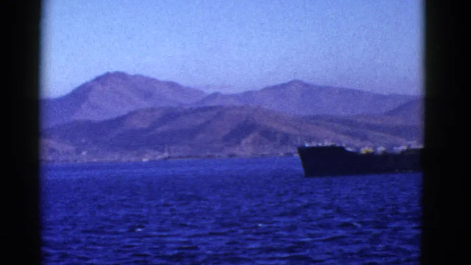 Only the right portion of the vessel remains in frame as the camera view opens onto blue water and a mountainous shoreline.