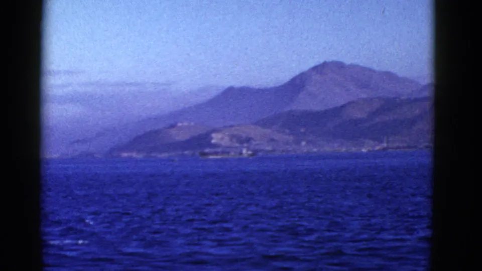 1950s San Francisco-area home-movie frame showing blue water, a low shoreline, and layered hills beyond.