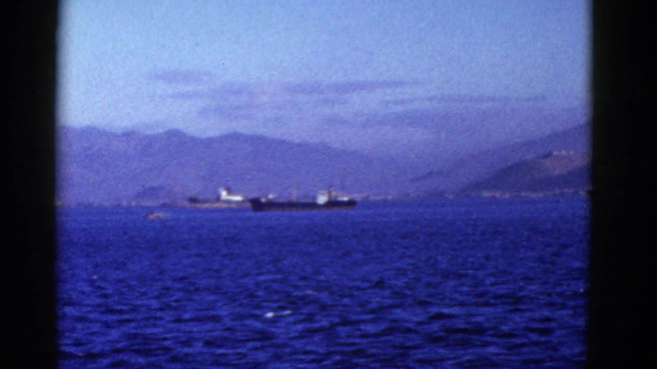 A dark-hulled ship crosses blue water in front of distant hills.