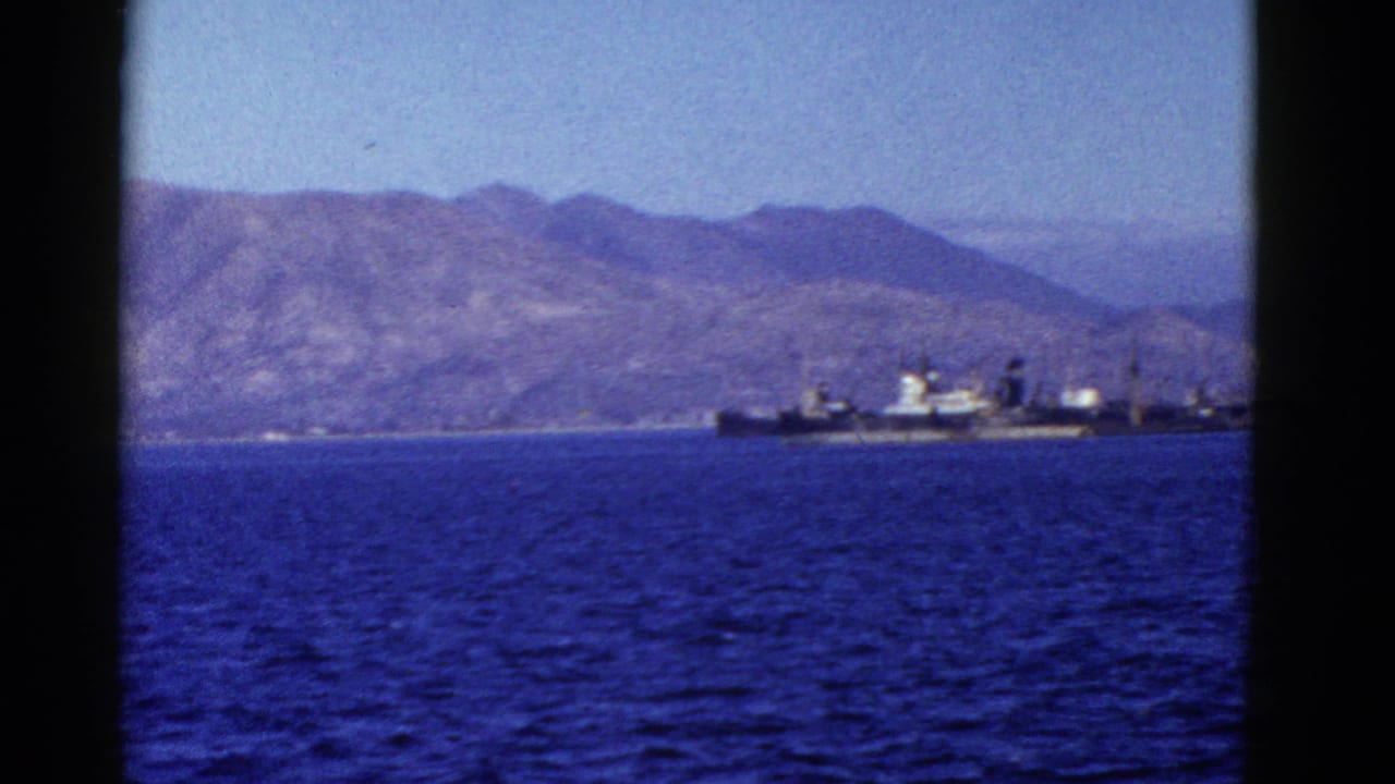 1950s San Francisco Bay home-movie frame showing a distant naval ship on blue water before dry rolling hills.